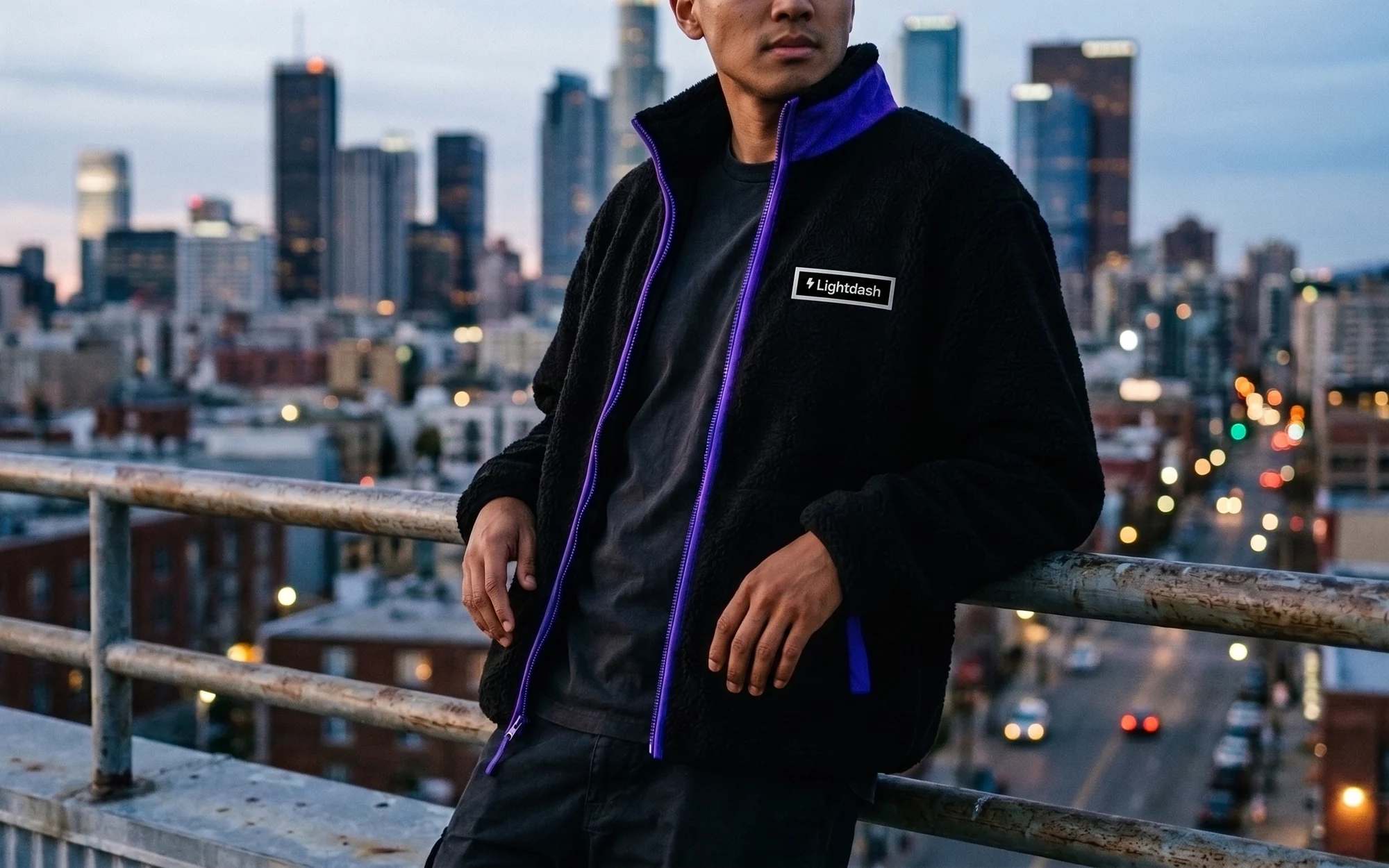 A man on a city rooftop at dusk in a black sherpa fleece with a purple zip and collar and a small Lightdash patch on the chest