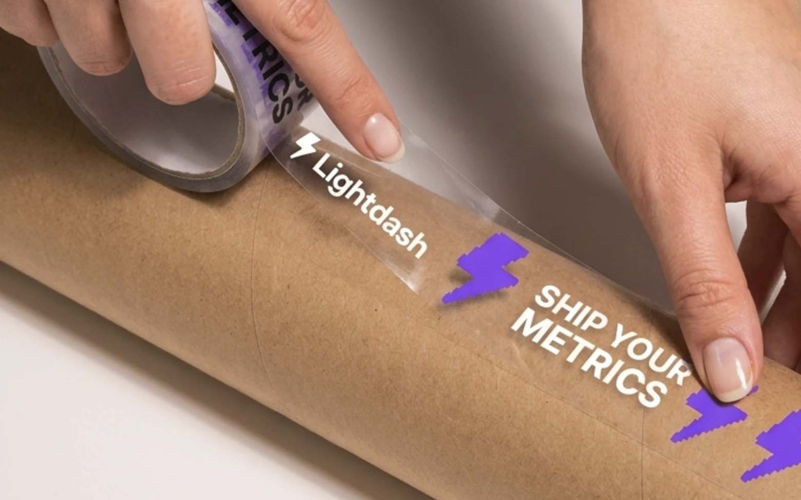 Two hands pressing clear Lightdash packing tape, printed with the wordmark, purple pixel bolts and Ship your metrics, onto a kraft mailing tube